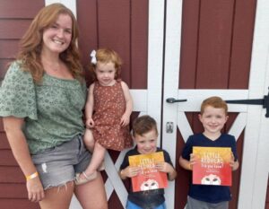 Redheaded family standing in front of a red barn on Redhead Celebration Day with two boys holding books for children with red heair